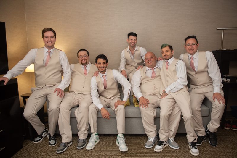 Seven men in matching cream vests, white shirts, and pink ties smile on a gray sofa, ready for a wedding.