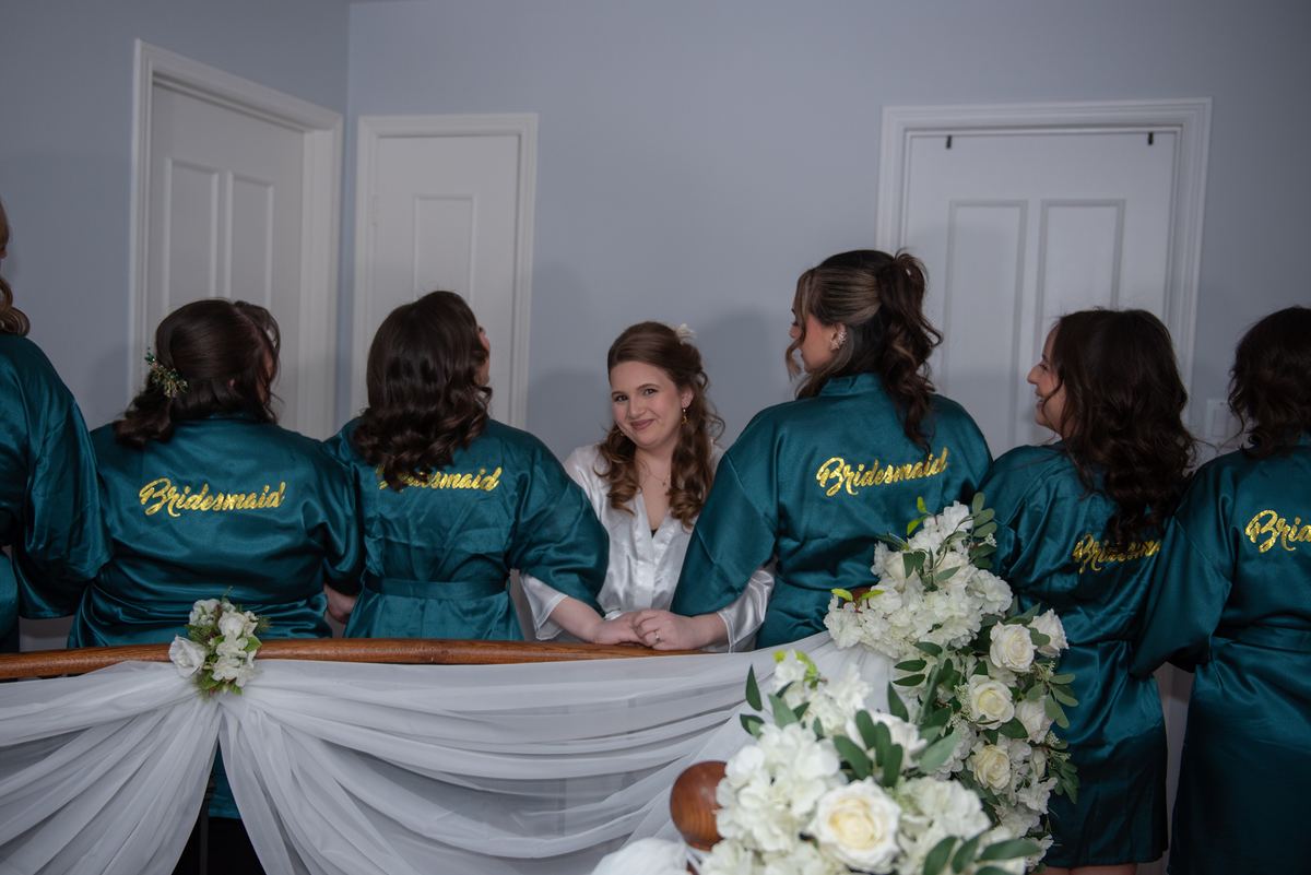 The bride in a white robe smiles with her bridesmaids in teal satin robes around a table with white floral arrangements.