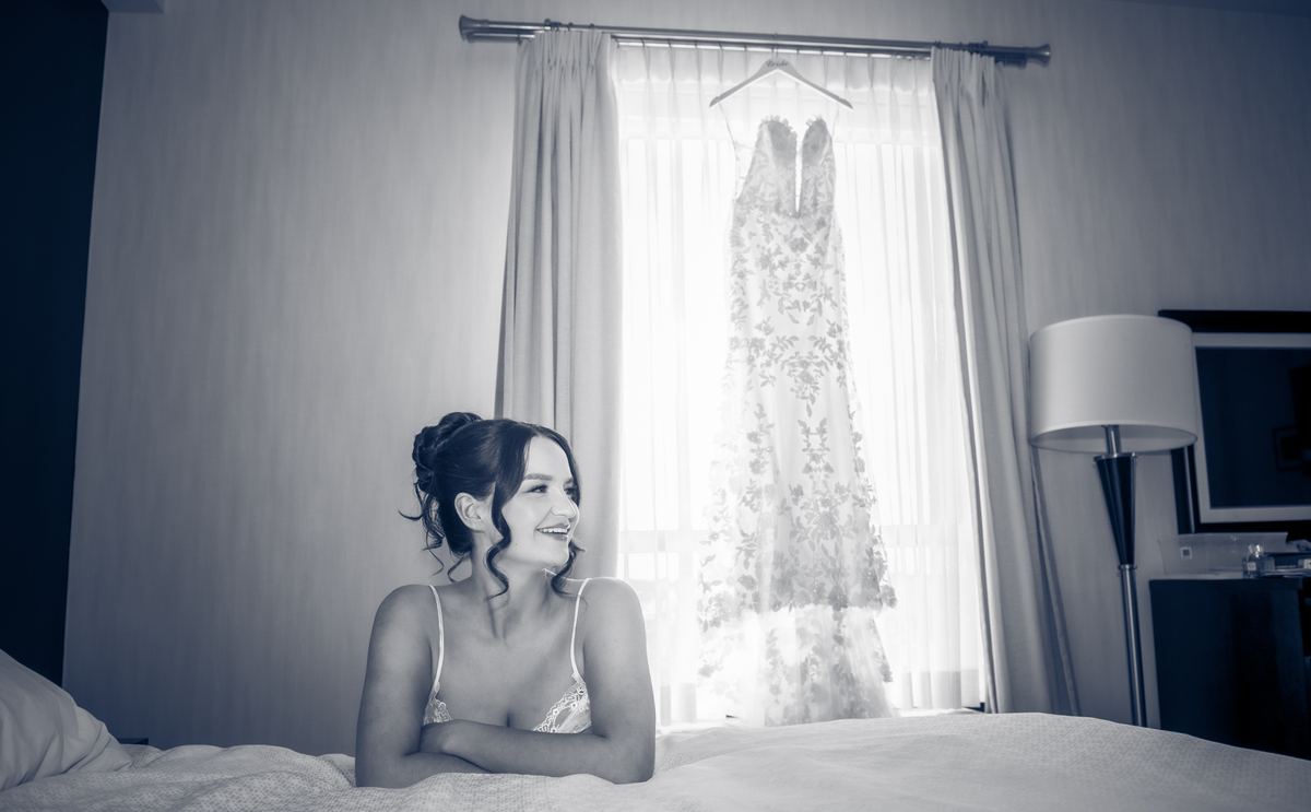 A bride smiles at her wedding gown hanging by the window as sunlight filters through sheer curtains.
