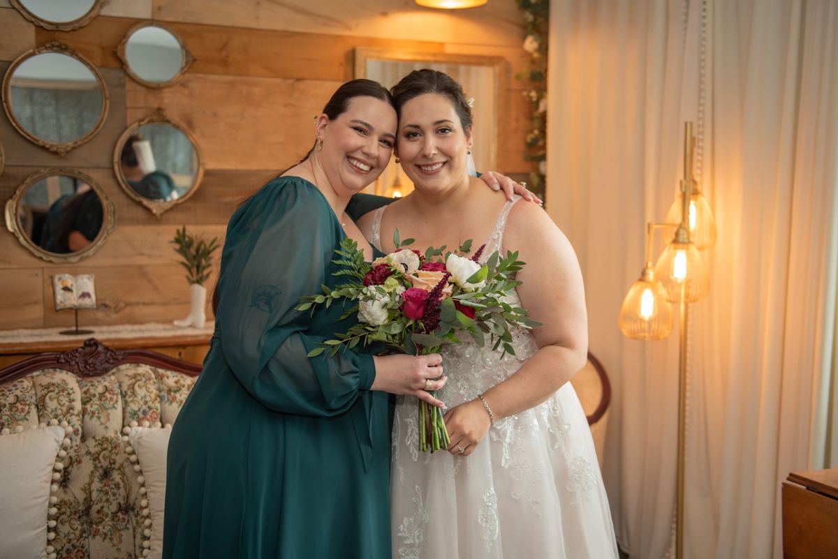 Two brides embrace warmly indoors, one holding a bouquet, in soft ambient light from pendant fixtures and mirrors.