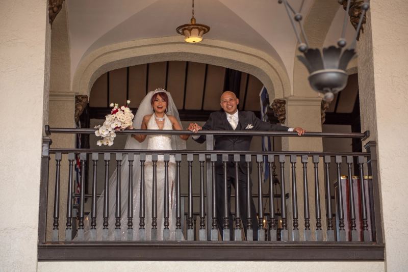A smiling bride and groom hold hands on an ornate balcony, illuminated by hanging fixtures and holding a bouquet.