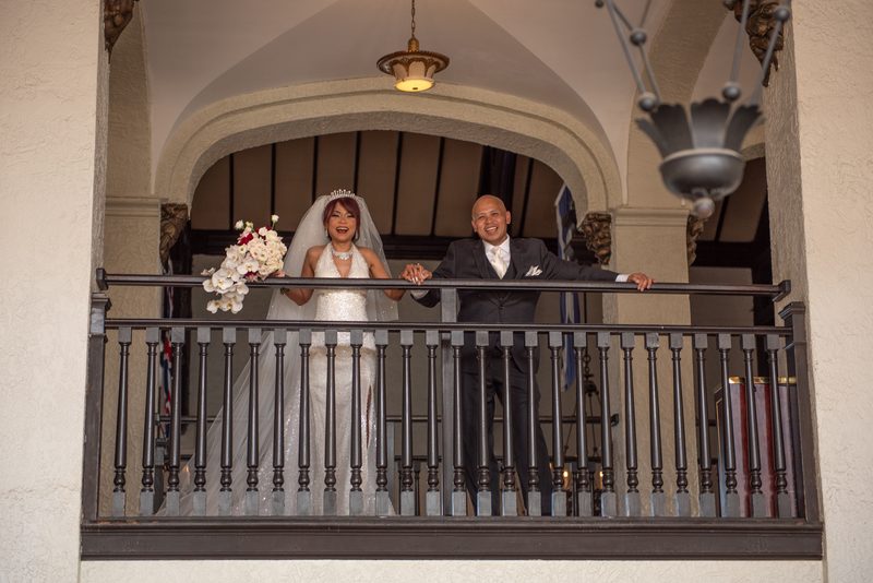 A smiling bride and groom hold hands on an ornate balcony, illuminated by hanging fixtures and holding a bouquet.