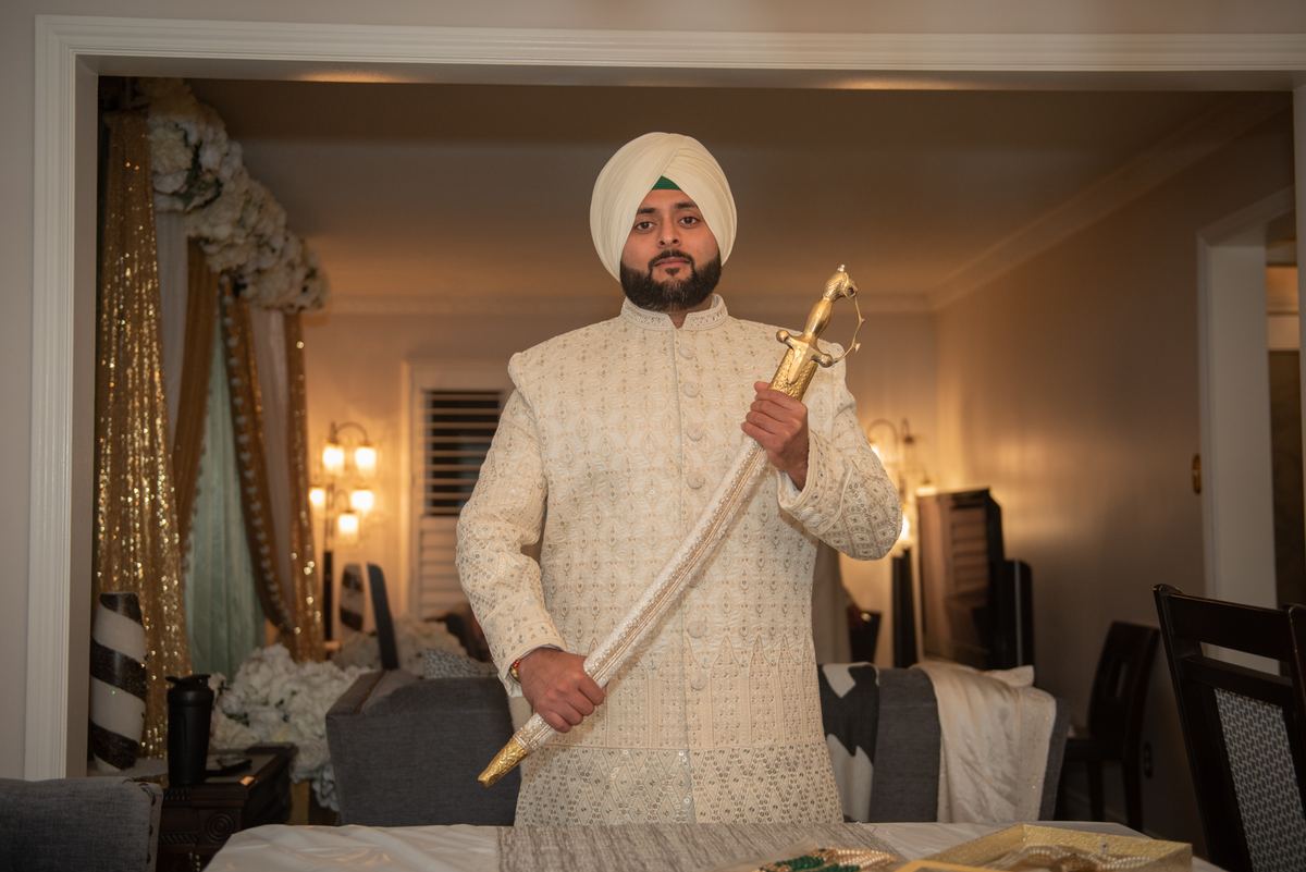 A Sikh groom stands quietly in a warmly lit indoor space, dressed in an ornate cream-colored sherwani and turban.