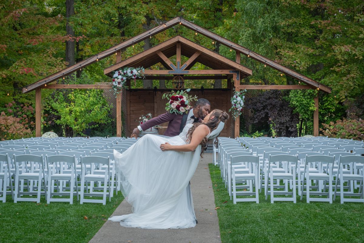 A newlywed couple kisses under a wooden arch, surrounded by autumn foliage and rows of white chairs.