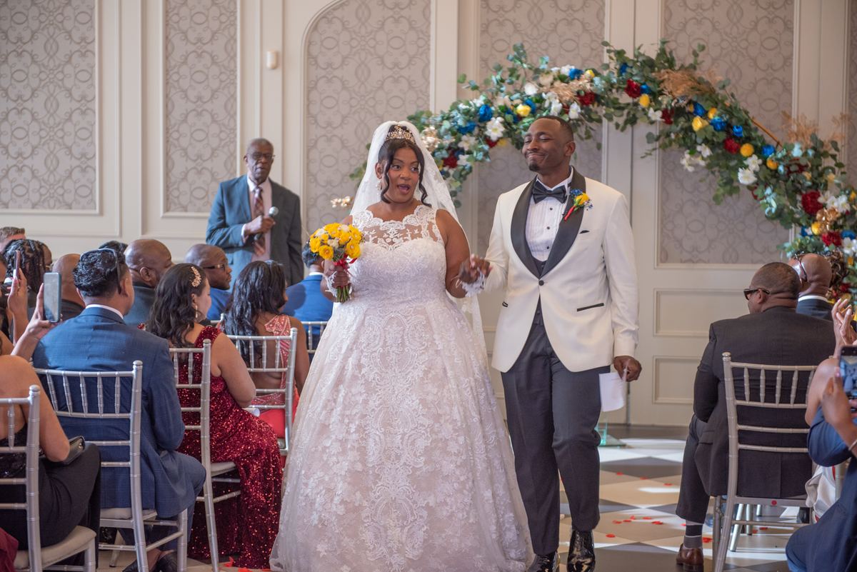 Bride and groom walk down the aisle during their wedding ceremony, surrounded by guests in silver chairs.