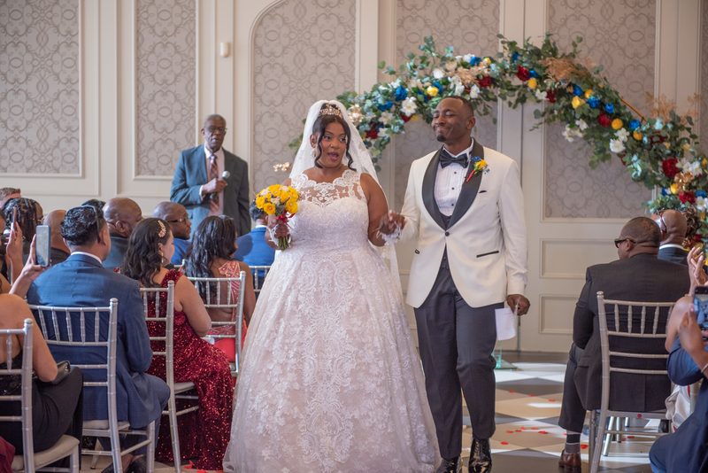 Bride and groom walk down the aisle during their wedding ceremony, surrounded by guests in silver chairs.