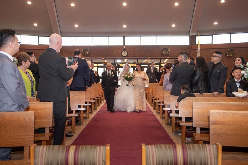 A bride in white and a bridesmaid in gold walk down the aisle of a modern church, illuminated by natural light.