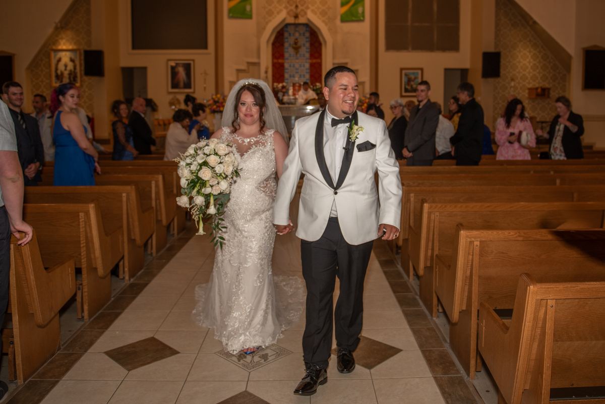 A bride in a lace gown and veil walks down the aisle with her groom in a white tuxedo toward the altar.