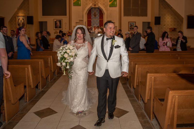 A bride in a lace gown and veil walks down the aisle with her groom in a white tuxedo toward the altar.