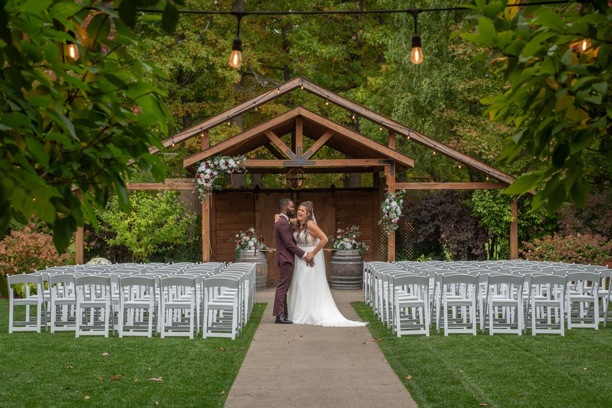 A newlywed couple embraces joyfully in front of white chairs under a wooden arbor decorated with flowers and Edison bulbs.