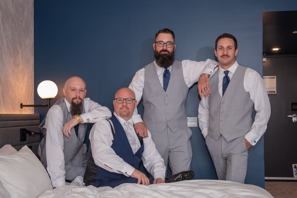 Four formally dressed men pose together in a room, one standing with arms around seated companions near a blue wall.