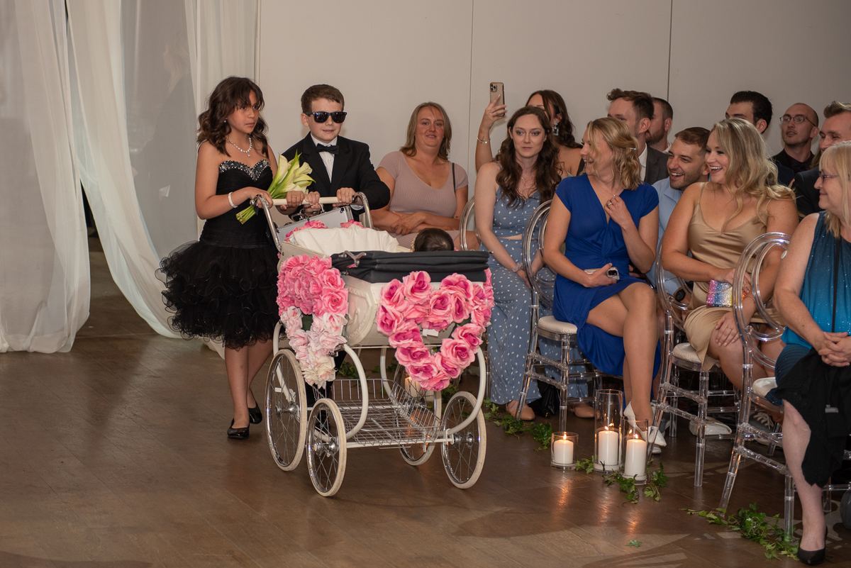 A girl and boy in formal wear walk down an aisle, pushing a pink-rosed carriage toward smiling guests.