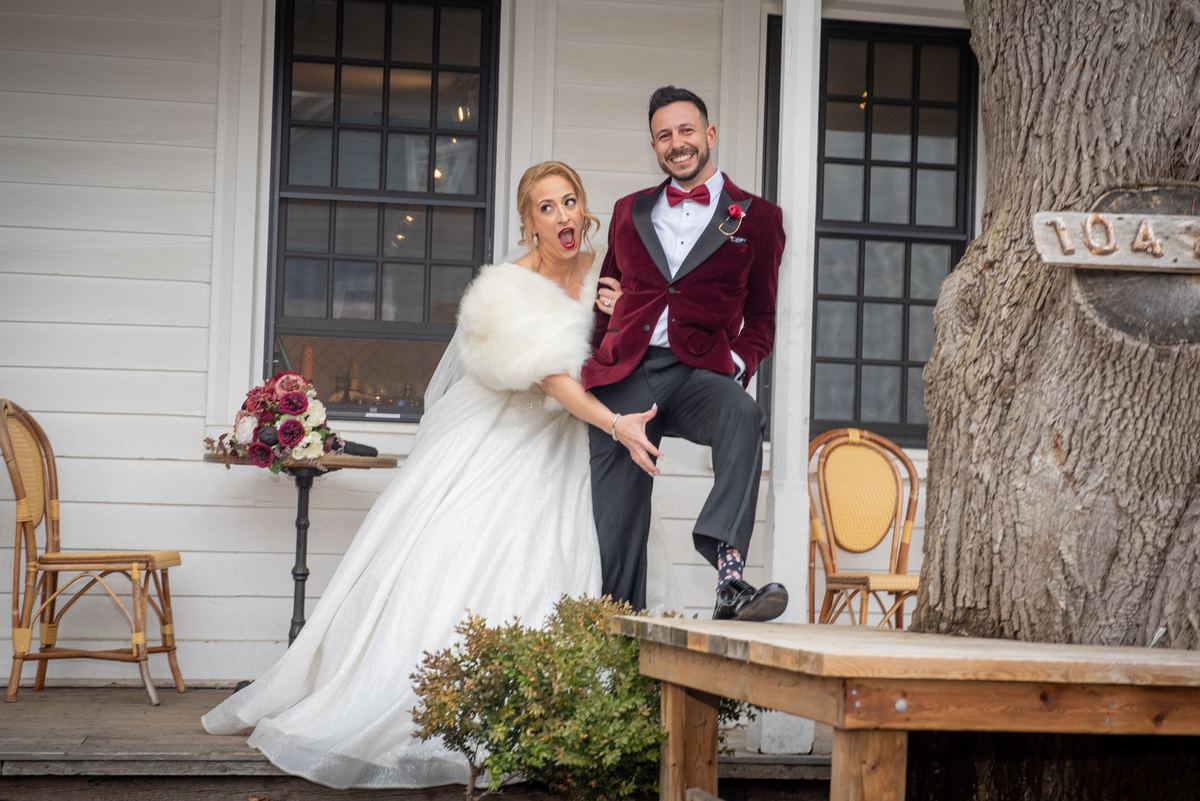 A bride and groom pose playfully on a porch, with the bride gasping dramatically and the groom smiling beside a wooden bench.