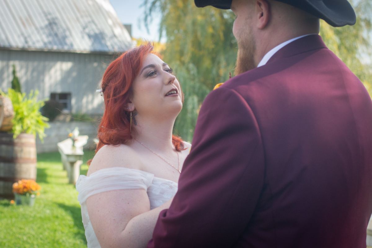 A red-haired bride in a white dress looks up at her groom in a burgundy suit during an outdoor wedding near a rustic barn.
