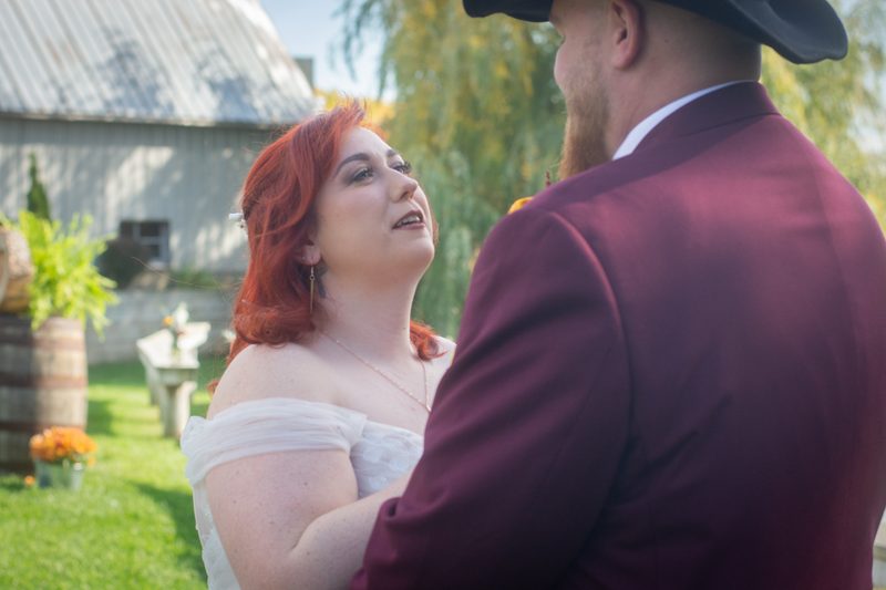 A red-haired bride in a white dress looks up at her groom in a burgundy suit during an outdoor wedding near a rustic barn.