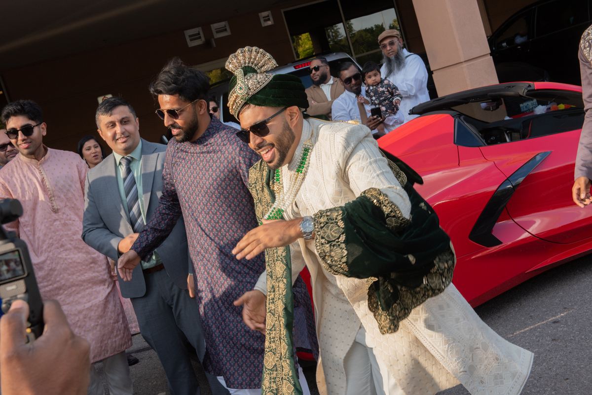 A groom in a white sherwani and gold turban smiles as friends lead him to a red convertible surrounded by guests.