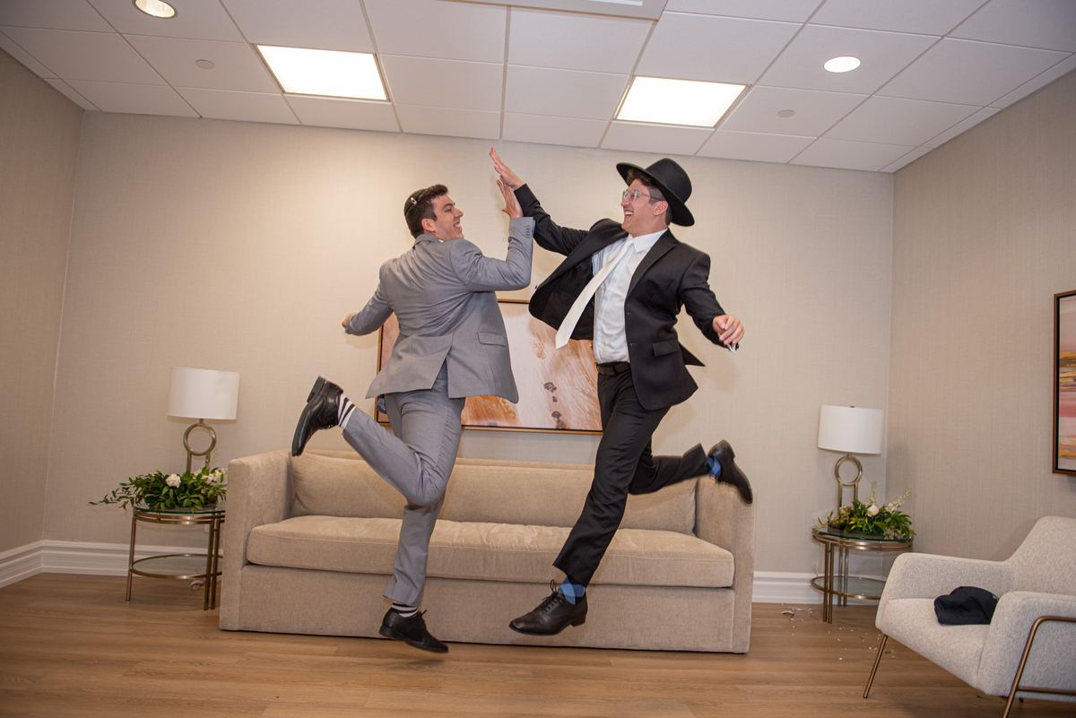Two men in formal suits joyfully jumping and high-fiving above a beige sofa in a modern living space, exuding celebration.