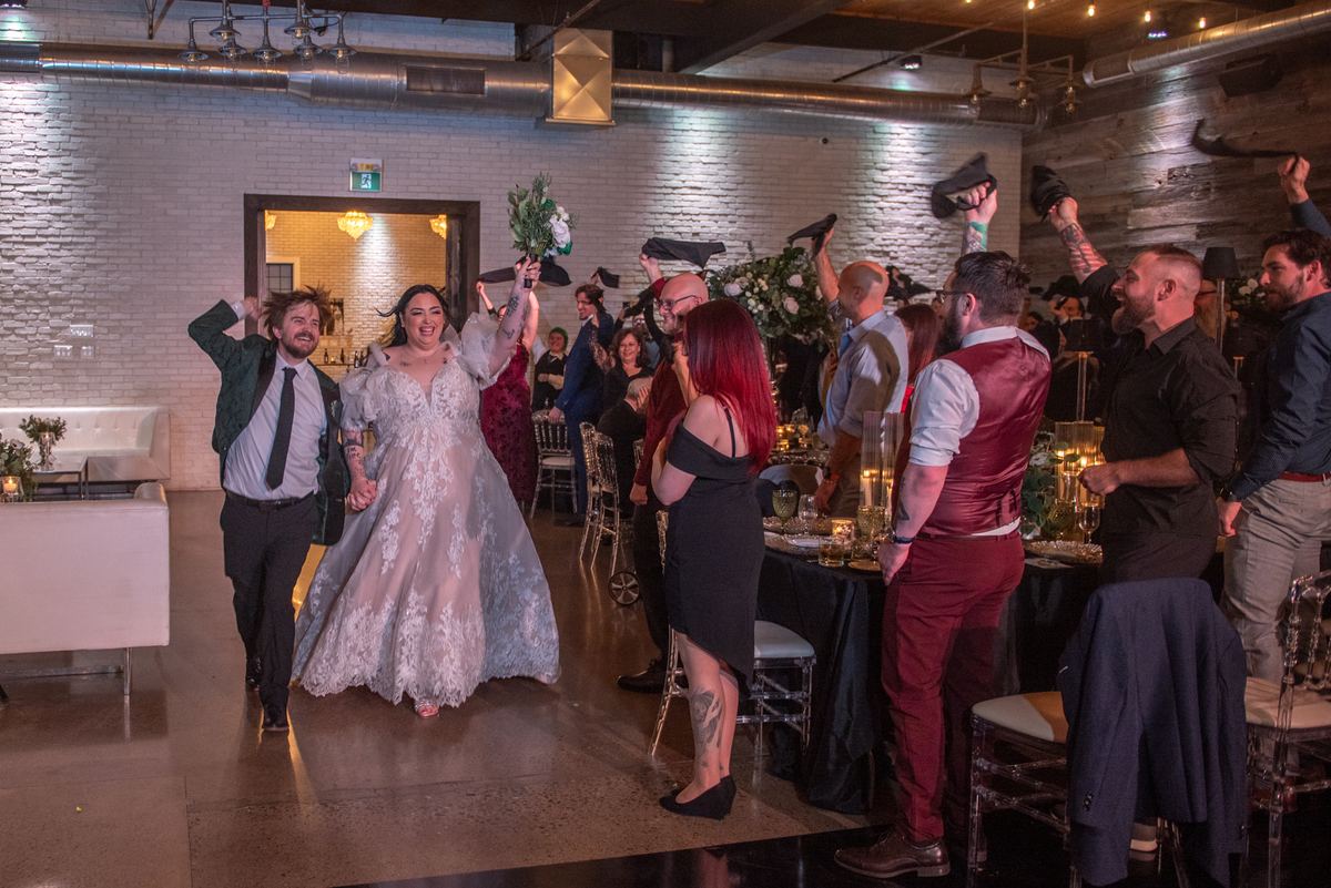 A smiling bride and groom walk through a cheering crowd at their reception, guests raising hats in celebration.