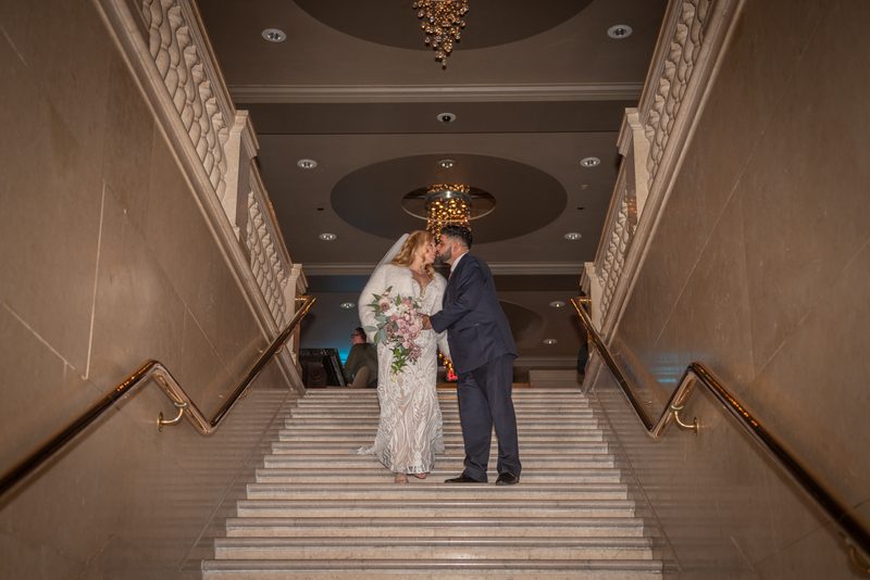 A bride and groom kiss tenderly on a grand marble staircase, surrounded by ornate chandeliers and warm lighting.