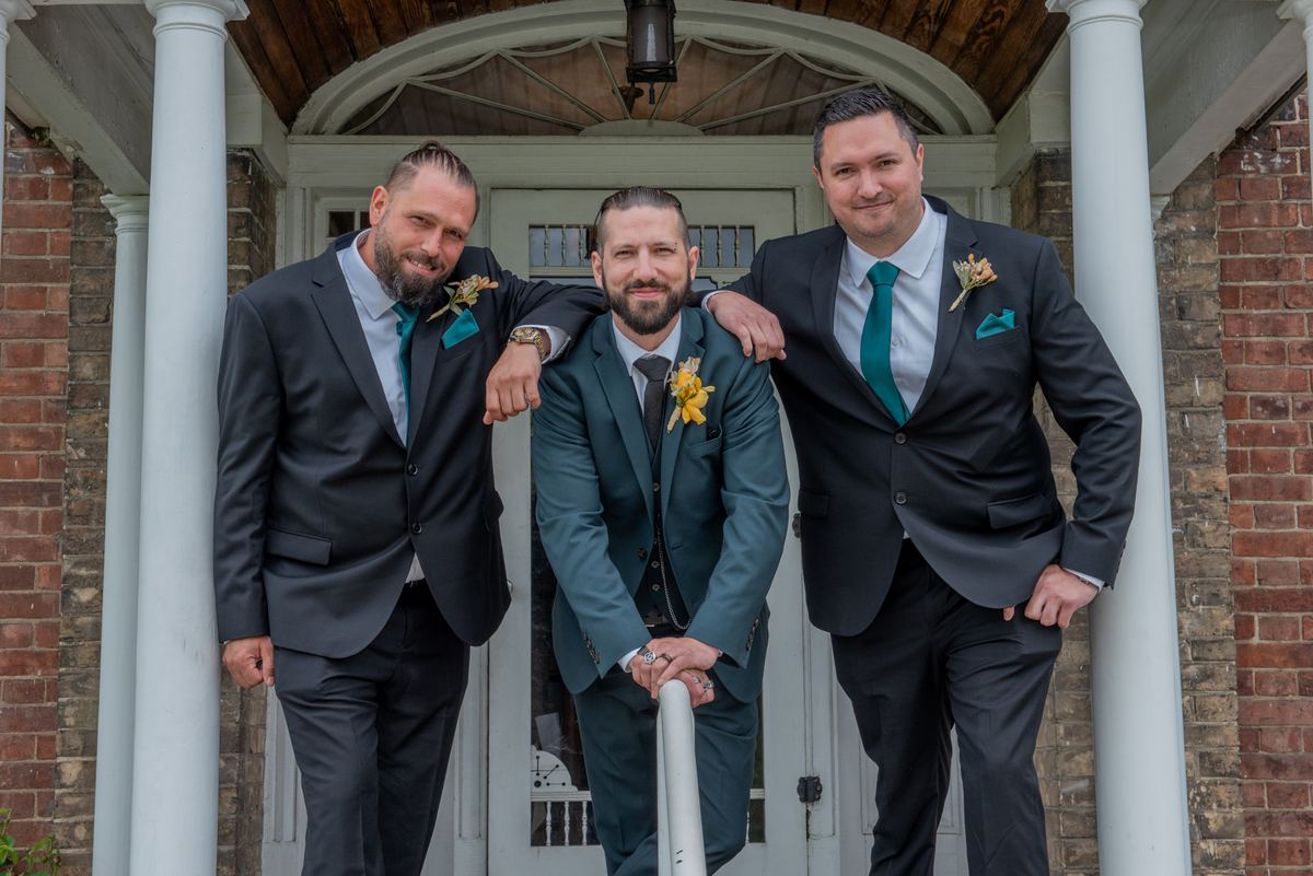 Three men in dark suits and teal ties stand confidently on a classic porch during a wedding celebration.