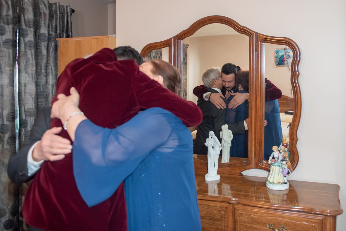 An intimate group hug in a warmly lit room, reflected in a triptych mirror, with small figurines on a dresser.