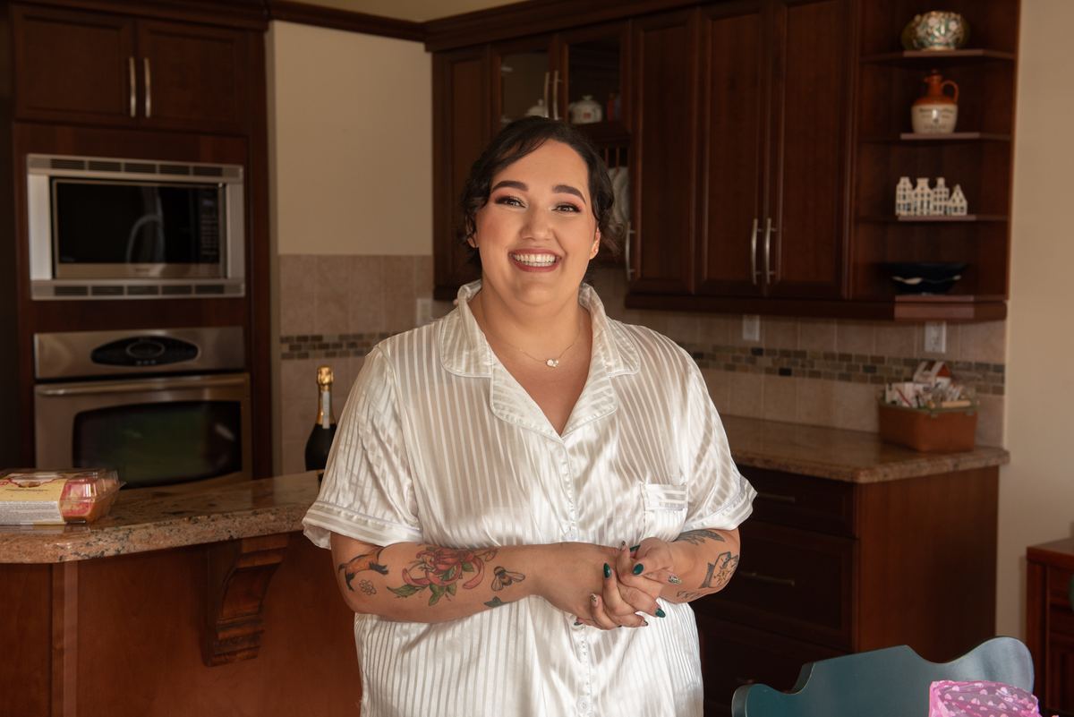 A joyful woman in white satin pajamas stands in her kitchen, smiling brightly at the camera next to a bottle of champagne.