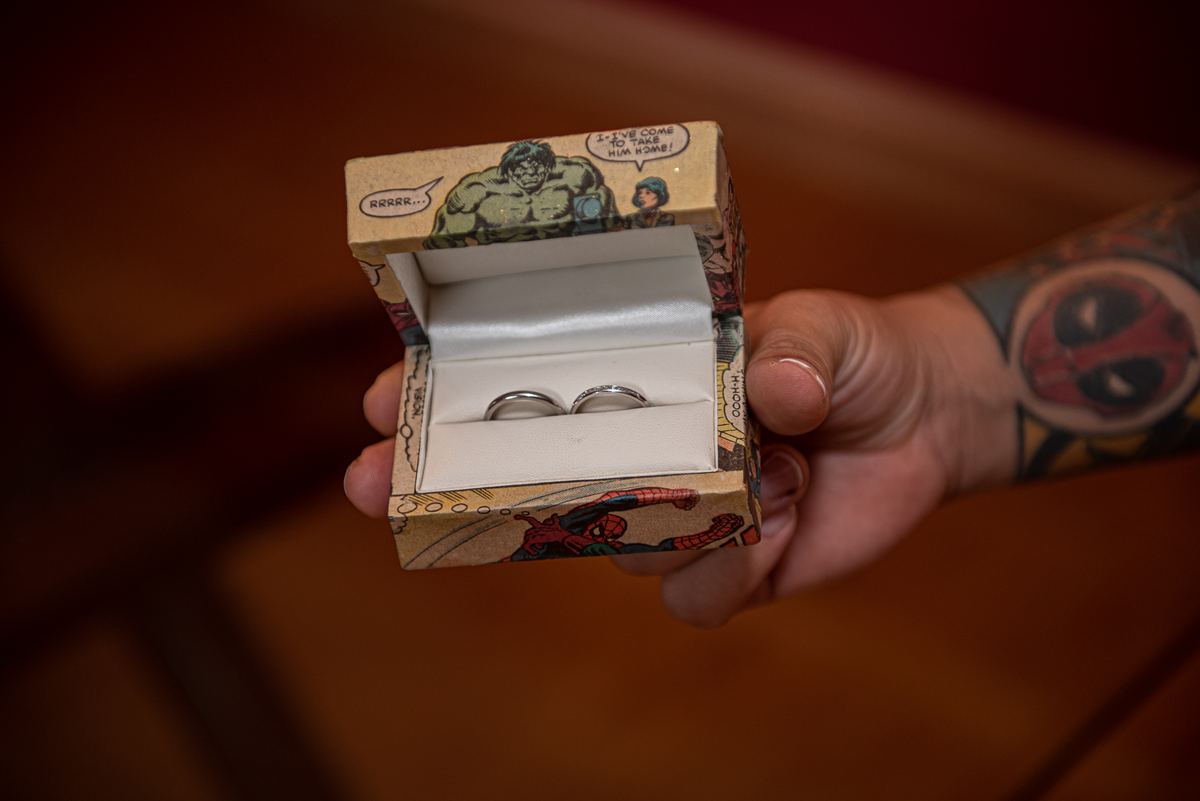 A hand holding open a comic book-style ring box revealing two wedding bands, with a Deadpool tattoo on the arm.