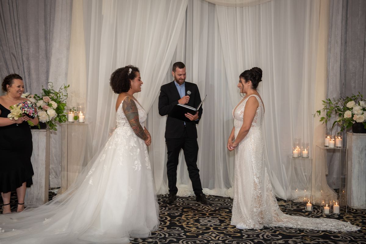 Two brides exchange vows in front of an officiant and a smiling bridesmaid, surrounded by soft candlelight.