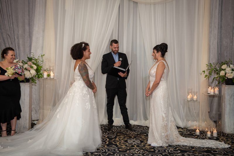 Two brides exchange vows in front of an officiant and a smiling bridesmaid, surrounded by soft candlelight.