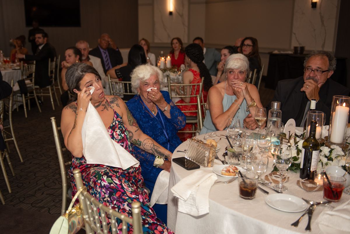 Guests at an elegant banquet react emotionally to a heartfelt speech, their faces illuminated by candlelight.
