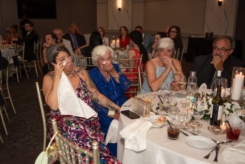 Guests at an elegant banquet react emotionally to a heartfelt speech, their faces illuminated by candlelight.