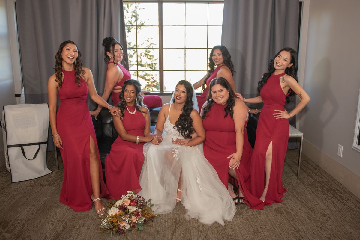 A bride in white sits with smiling bridesmaids in red dresses, surrounded by soft natural light and a floral bouquet.