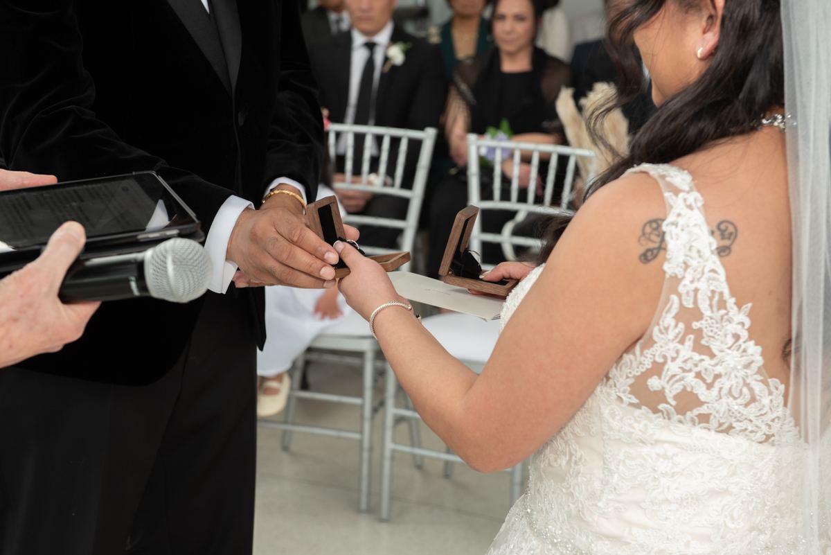 Bride and groom exchange rings under officiant's guidance, with guests in formal attire visible behind them.