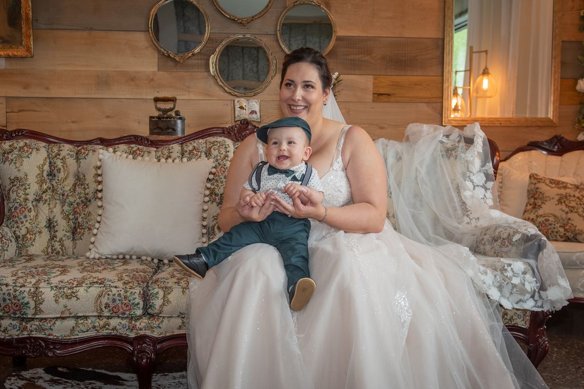 A bride in wedding attire sits on an ornate floral sofa with a toddler dressed formally, surrounded by rustic decor.