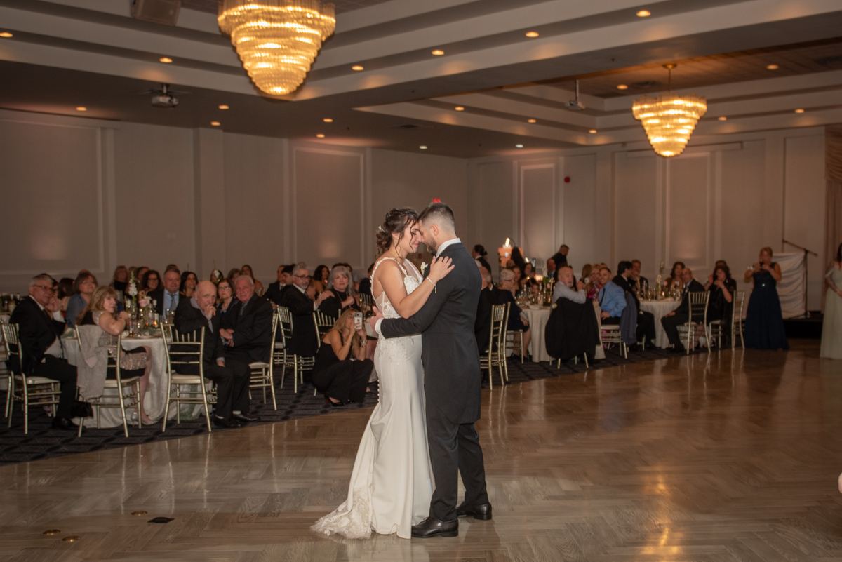 A bride and groom dance intimately at their reception, surrounded by smiling guests and illuminated by chandeliers.