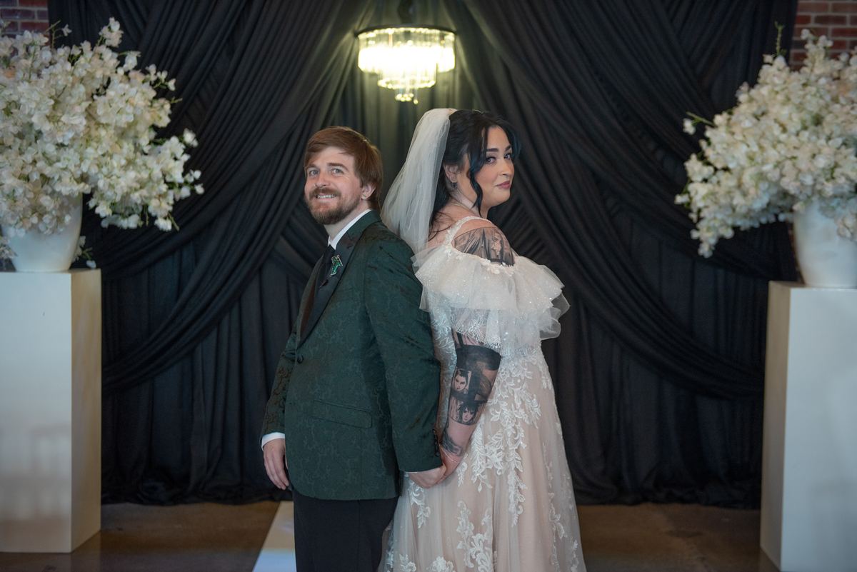 A bride and groom in a stylized setting, back-to-back, smiling at the camera with soft lighting from a chandelier.