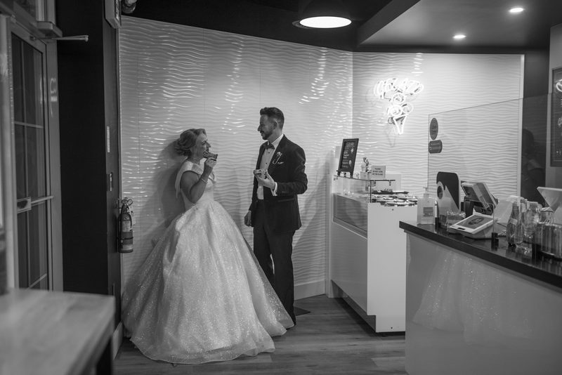 A bride and groom laugh together near a dessert counter, holding small bites of food under soft neon lights.