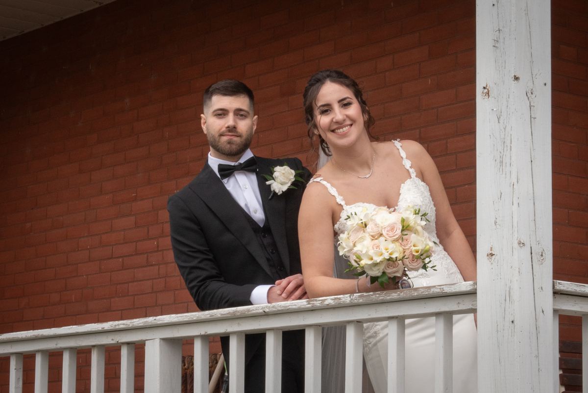 A smiling bride and groom stand on a porch railing, the bride holding roses against a red brick wall.