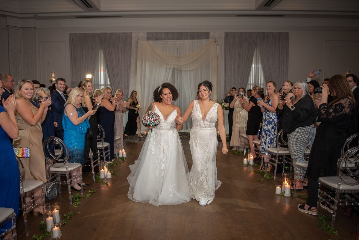 Two brides walk hand-in-hand down the candlelit aisle, smiling and laughing as guests applaud, illuminated by warm candles.