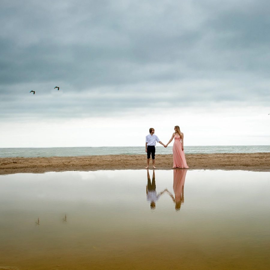 Two small figures hold hands on a wide, pale beach, mirrored in the flat water beneath an overcast sky.