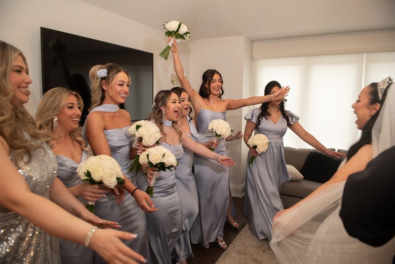 Bridesmaids in silver-gray gowns cheerfully welcome the bride with outstretched arms and raised bouquet.
