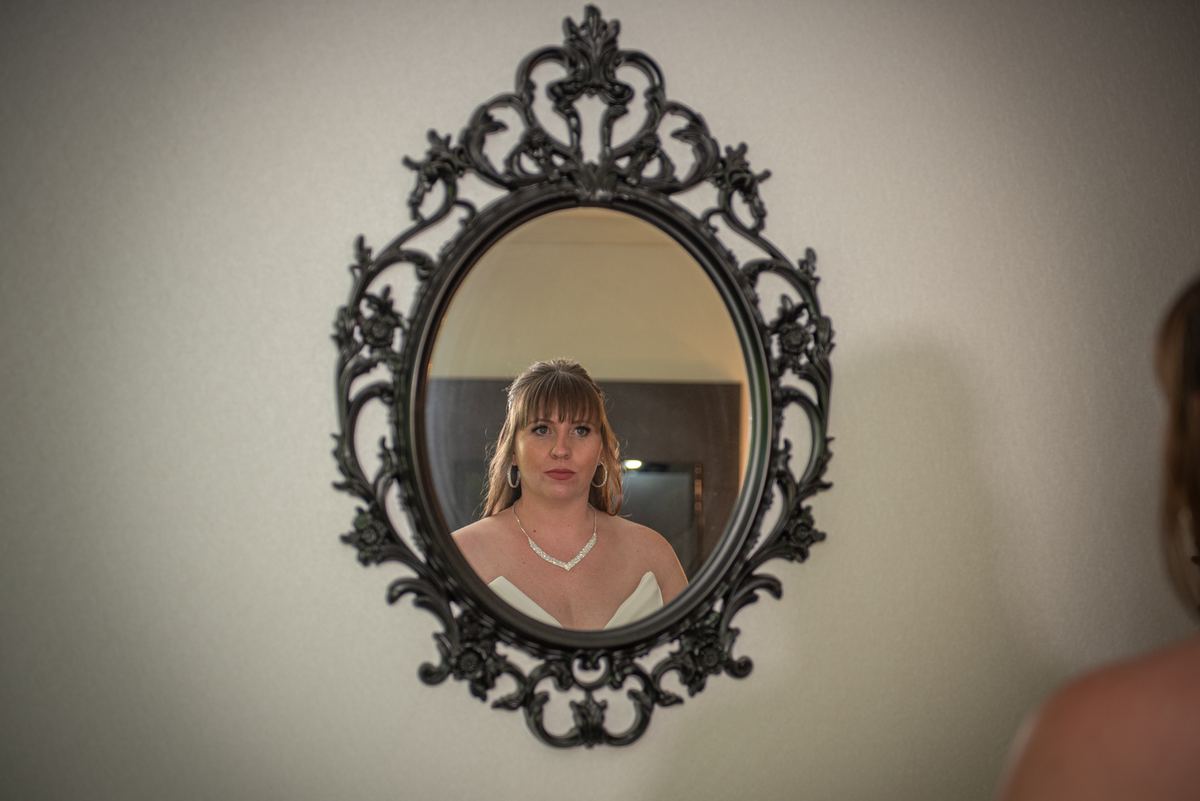 A bride stands before an ornate mirror, gazing thoughtfully at her reflection in the warm, soft light.