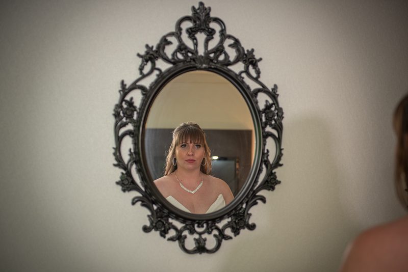 A bride stands before an ornate mirror, gazing thoughtfully at her reflection in the warm, soft light.
