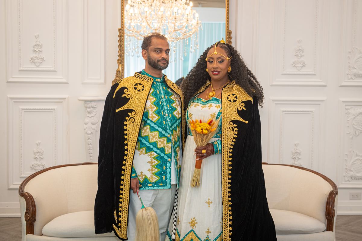 A formally dressed couple in traditional attire stands in an ornate room with a chandelier visible behind them.