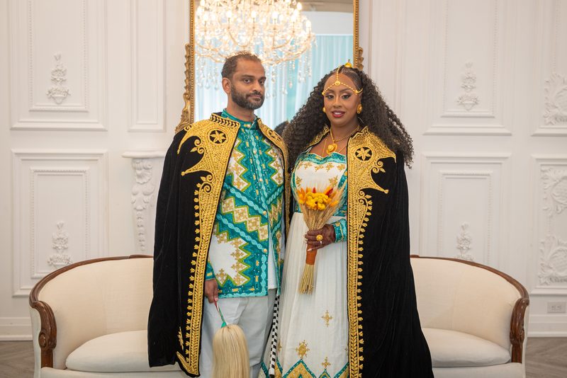 A formally dressed couple in traditional attire stands in an ornate room with a chandelier visible behind them.