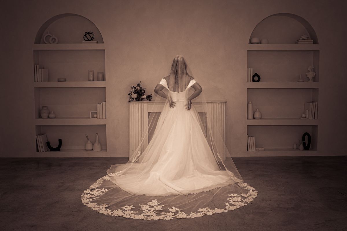 A bride in a white gown and veil stands with hands on hips before arched shelves, bathed in soft light.