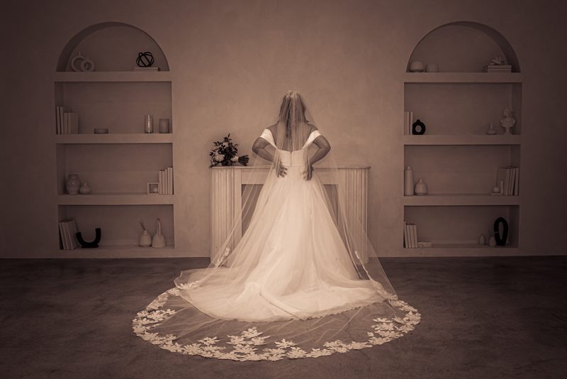 A bride in a white gown and veil stands with hands on hips before arched shelves, bathed in soft light.