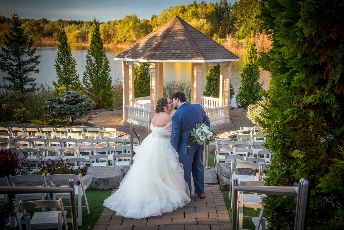 Morgan and Aaron share a tender kiss at their wedding ceremony, framed by a picturesque gazebo overlooking a tranquil lake.