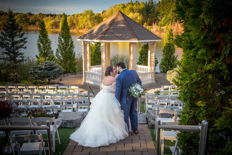 Morgan and Aaron share a tender kiss at their wedding ceremony, framed by a picturesque gazebo overlooking a tranquil lake.
