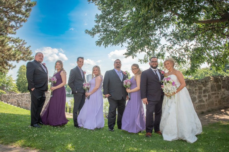 A bridal party poses happily outdoors under a partly cloudy sky with dappled sunlight through trees.